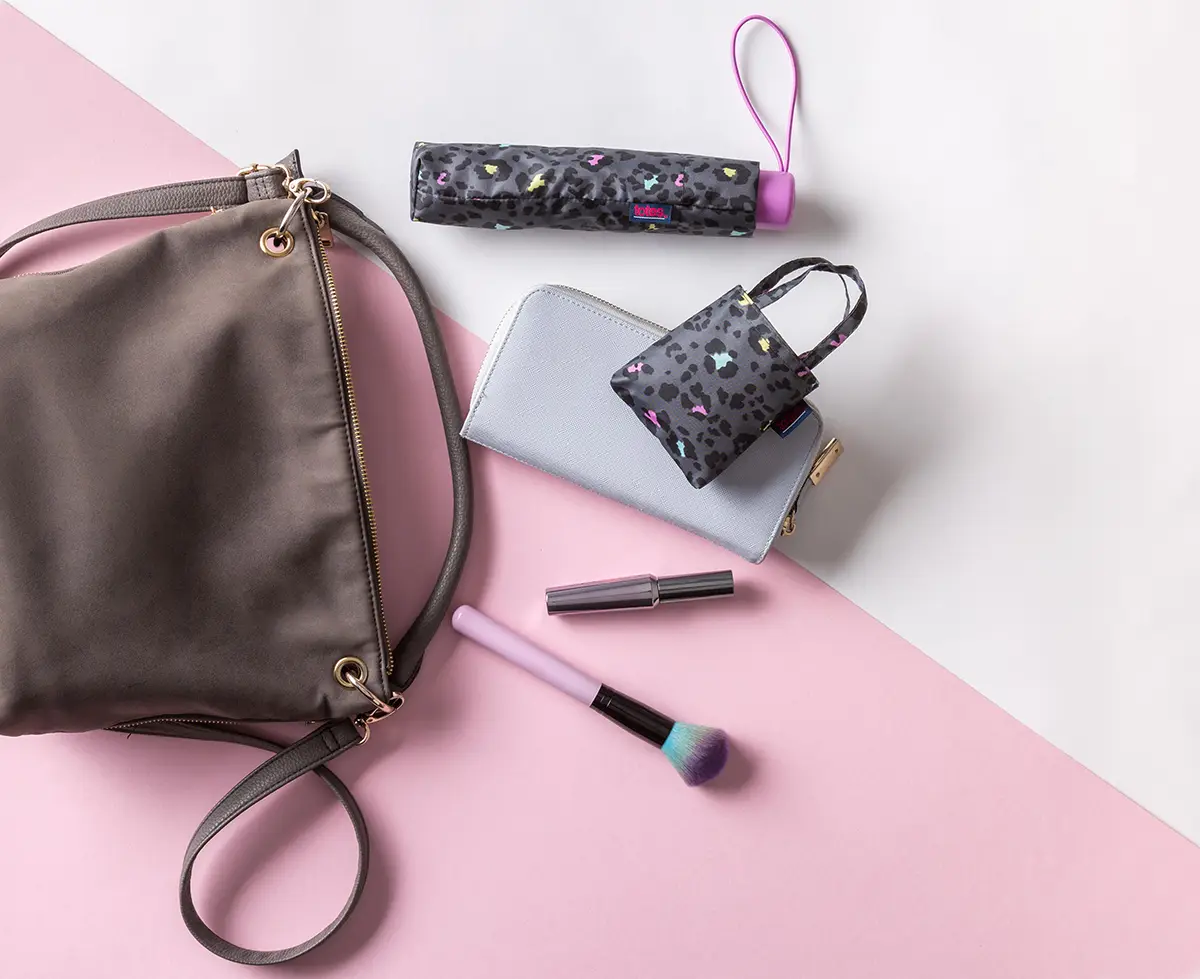 An image of some Totes umbrella products laid out next to a brown bag, on a pink and white background